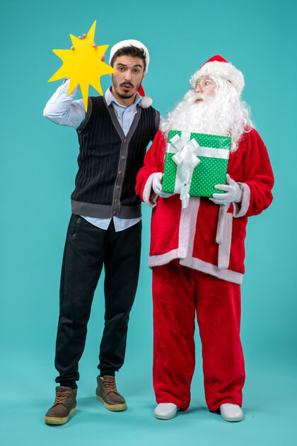 圣诞前视图：圣诞老人和手持蓝色背景黄色标志的年轻男性举行年轻人帽子