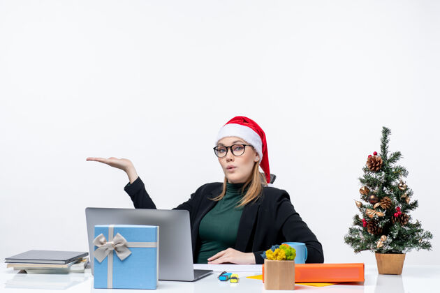 礼物震惊的年轻女子戴着圣诞老人的帽子和眼镜坐在一张桌子旁 桌子上有一棵圣诞树和一份白色背景的礼物女人工作专业