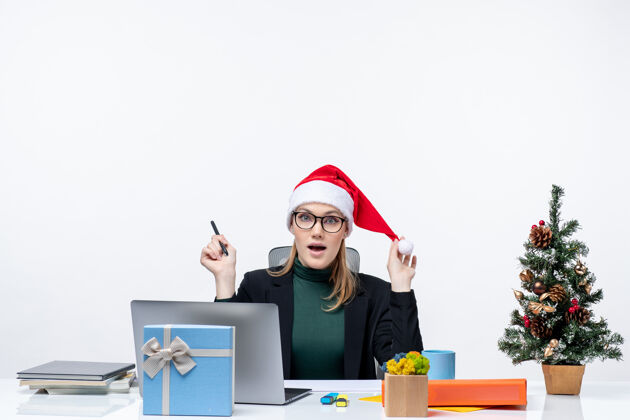 工作新年的气氛 好奇的金发女人 戴着圣诞老人的帽子 坐在一张桌子旁 桌子上有一棵圣诞树和一份白色背景的礼物女人笔记本电脑圣诞节