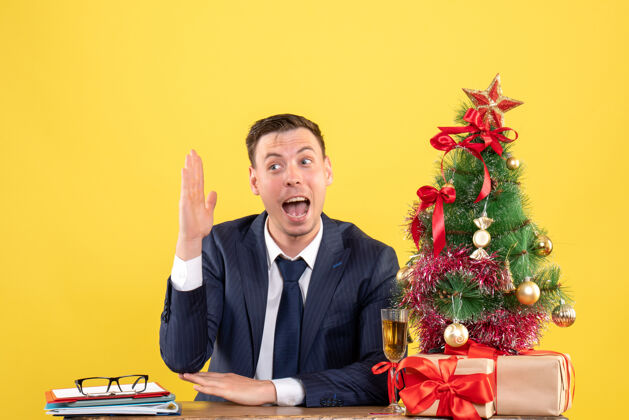 男人年轻人举起他的手坐在圣诞树旁的桌子上 用黄色的礼物桌子商人年轻人