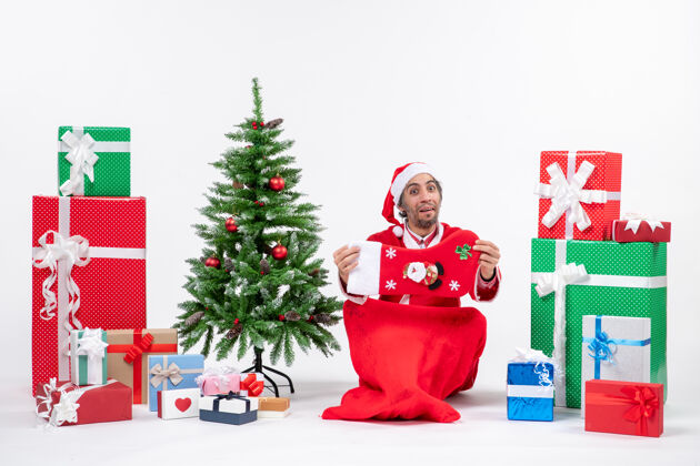袜子新年气氛有趣积极的圣诞老人坐在地上 拿着圣诞袜子附近的礼物和装饰圣诞树上的白色背景拿着圣诞老人礼物