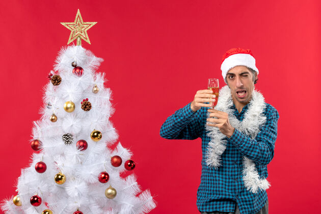 玻璃圣诞节心情与有趣的年轻人圣诞老人帽子在一个蓝色条纹衬衫举起一杯葡萄酒附近圣诞树人新前夜