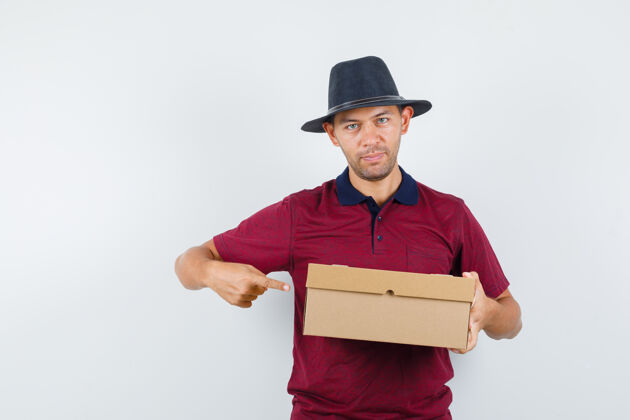 盒子年轻男子穿着红衬衫 戴着黑帽子指着盒子 看上去很感兴趣 前视图帽子帅气肖像