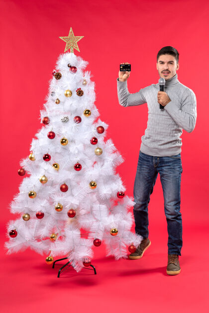 美丽圣诞节的气氛与自信的家伙穿着牛仔裤站在装饰圣诞树附近 手持麦克风和拍摄视频的每个人红色自信圣诞
