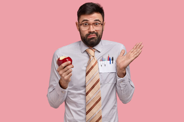 正式莫名其妙的不满满脸胡须的男人摊开手 拿着红苹果 戴着眼镜和正装 感到疑惑问题工人咬人