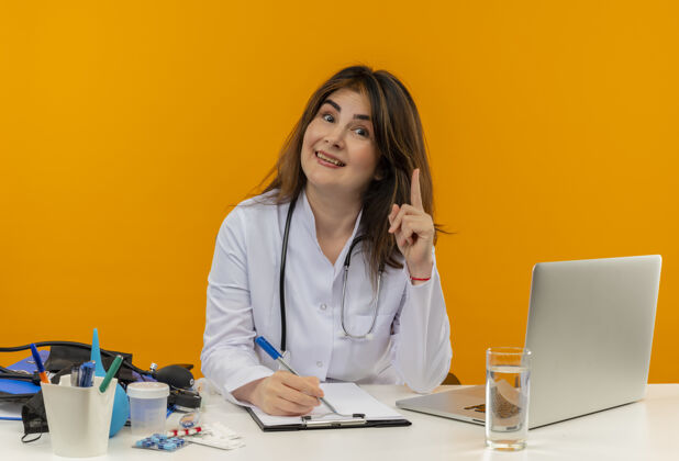点高兴的中年女医生穿着医用长袍带着听诊器坐在办公桌上用笔记本电脑带着医用工具写在剪贴板上 指着隔离的橘黄色墙上有复印空间中年书写坐