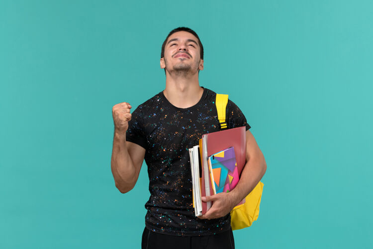 男性前视图中的男学生在黑暗的t恤黄色背包举行文案和文件在蓝色的墙上学生服装服装
