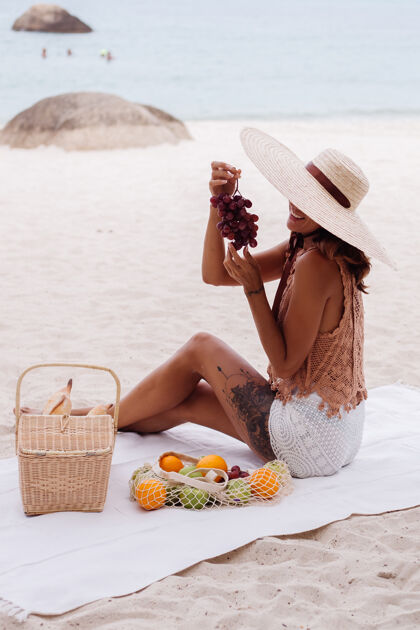 快乐年轻漂亮的白种人 皮肤黝黑 穿着针织衣服 戴着帽子在沙滩上上衣套装手提包