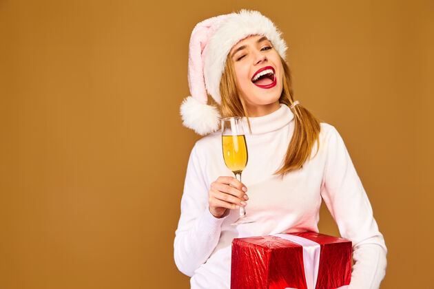 年轻戴着圣诞帽和大礼盒的模特在金墙上喝香槟友好帽子时尚