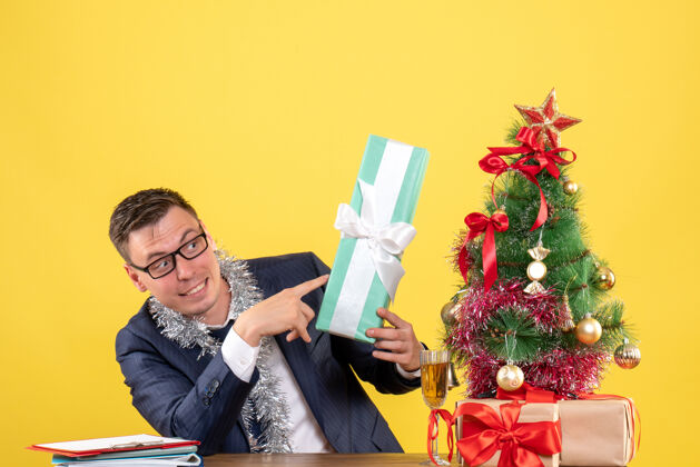 黄色年轻人指着坐在圣诞树旁桌上的礼物 把礼物放在黄色的桌子上圣诞树桌子年轻人