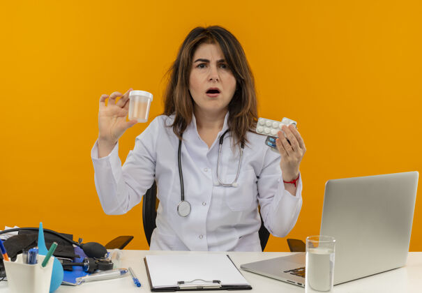 中年困惑的中年女医生穿着医用长袍 手持听诊器坐在办公桌旁 手提电脑上拿着医疗工具 拿着空罐子和药片 孤零零地贴在橙色的墙上医疗女性工作