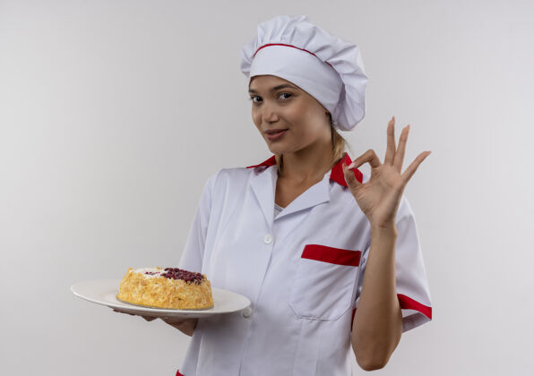 厨师高兴的年轻厨师女穿着厨师制服 拿着蛋糕在盘子上 在隔离的白色墙壁上显示okey手势与复制空间高兴好的厨师