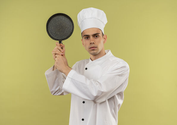 肩膀年轻的男厨师穿着厨师制服 肩上扛着煎锅站在隔离的绿墙上穿着厨师制服