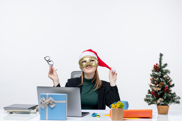 帽子圣诞节的心情与梦幻的年轻女子玩圣诞老人的帽子 戴着眼镜和面具坐在一张白色背景的桌子上笔记本电脑坐着工作