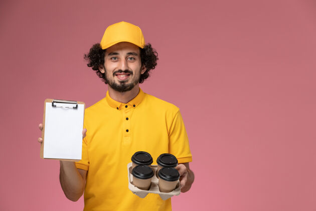 手持正面图：身穿黄色制服的男信使手拿棕色的咖啡杯 粉色墙上放着记事本剪贴板记事本帽子