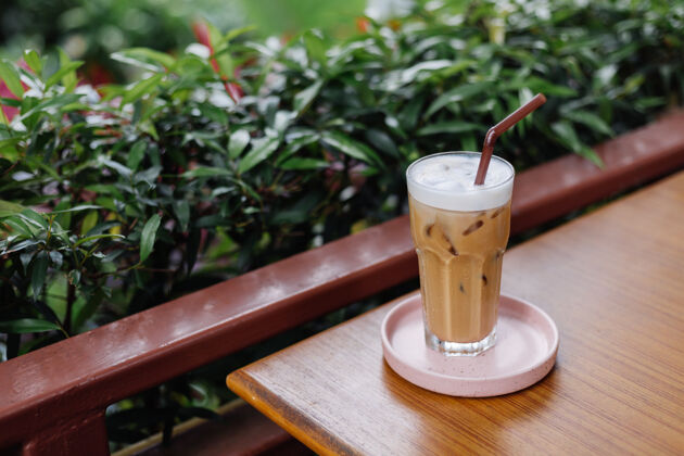 棕色在绿色灌木丛的夏日咖啡馆里 粉红色的架子上放着一杯冰拿铁早晨奶油下午茶