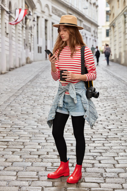 手机时尚女人的垂直镜头戴帽子 条纹套头衫 牛仔裤短裤和橡胶红靴子 手持蜂窝阅读饮料城市