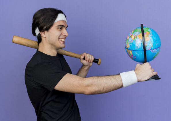 穿着年轻的运动型男子穿着运动服 头戴棒球棒和地球仪 愉快地微笑着站着地球仪抱着