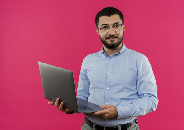 胡子戴着眼镜 穿着蓝色衬衫 留着胡子的年轻人拿着笔记本电脑 自信地微笑着自信衬衫蓝色