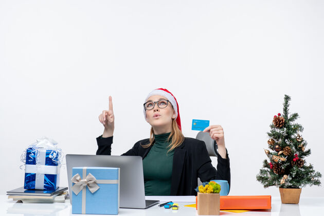 指着一位戴着圣诞老人帽 戴着眼镜 手拿着银行卡 坐在桌旁的迷人女人指着办公室的上方女人圣诞老人银行