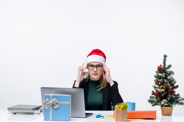 帽子新年心情坚定的女人戴着圣诞老人的帽子坐在一张桌子旁 桌子上有一棵圣诞树和一份白色背景的礼物电脑笔记本电脑礼物