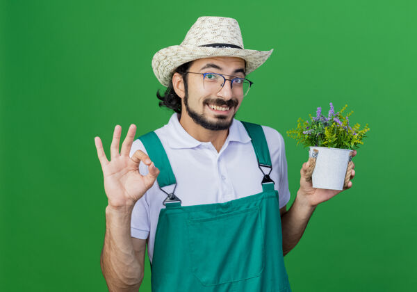 显示年轻的留着胡须的园丁 穿着连体衣 戴着帽子 手里拿着盆栽植物 微笑着表示同意站着微笑植物