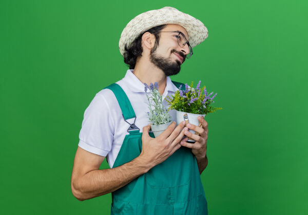 男人年轻的留着胡须的园丁 穿着连体衣 戴着帽子 手里拿着盆栽植物 感受着积极的情绪植物穿着连身衣