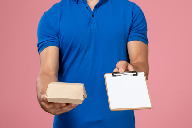 男性正面图身穿蓝色制服披肩的年轻男性快递员拿着小快递食品包和浅粉色墙上的记事本食物送货工作
