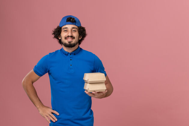 人正面图：身穿蓝色制服 披风的男性快递员在粉红色的墙上拿着小包裹食物快递制服