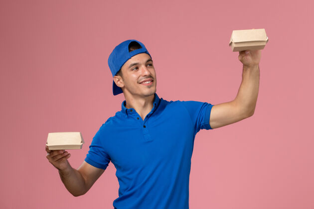 送货正面图：身穿蓝色制服披肩的年轻男性快递员在粉红色的墙上拿着小包裹服务工作年轻人