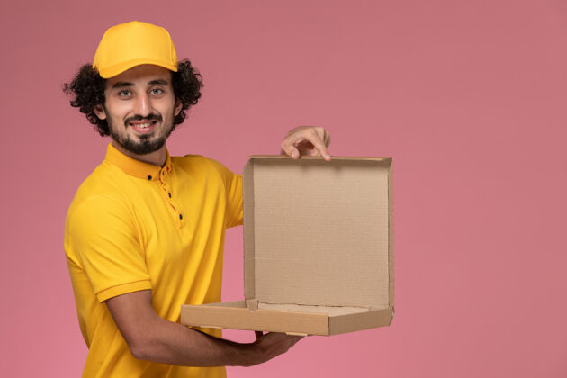食物正面图身穿黄色制服的男快递员拿着淡粉色墙上的空食品盒视图工作工作