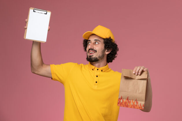 人正面图：身穿黄色制服的男快递员拿着食品包 粉色墙上放着记事本快乐黄色男快递