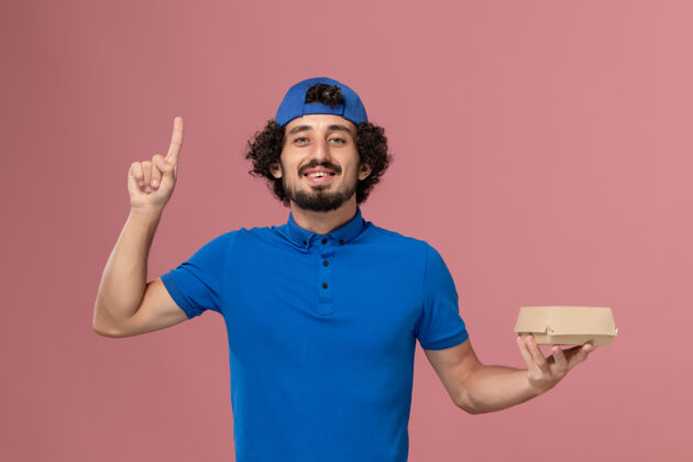 服务正面图：身穿蓝色制服和披风的男快递员举着一个小小的快递食品包 手指举在粉红色的墙上手指包裹男性
