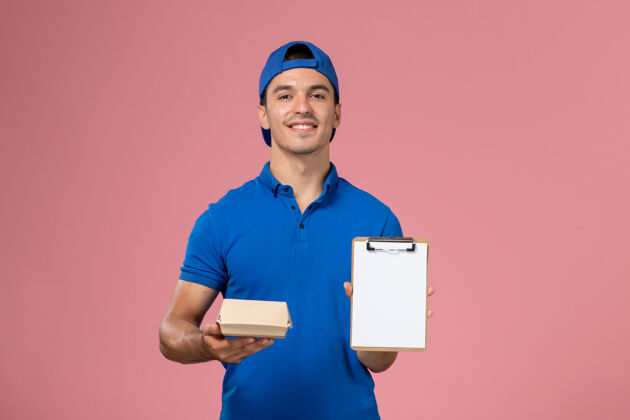 人正面图身穿蓝色制服披肩的年轻男性快递员拿着小快递食品包和浅粉色墙上的记事本年轻男性快递工作汉堡