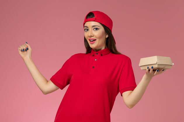 视图正面图：身穿红色制服和斗篷的年轻女快递员 手上拿着一个小小的快递食品包 放在粉红色的墙上粉色食物女人