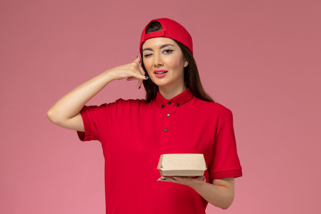 微笑正面图：身穿红色制服和斗篷的年轻女快递员 手上拿着一个小小的快递食品包 放在粉红色的墙上成人女人人