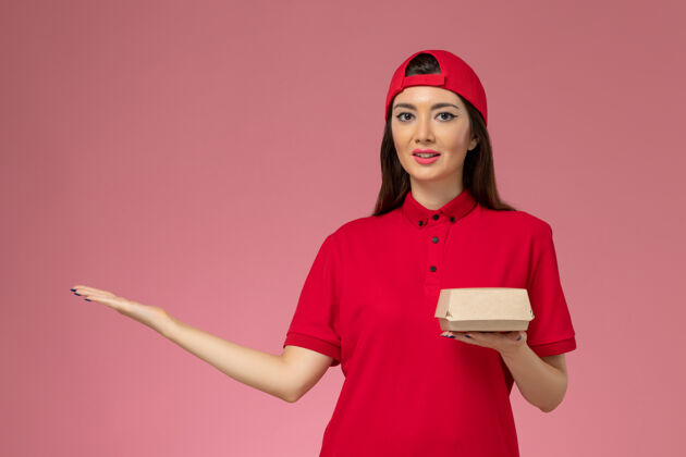 女人正面图：身穿红色制服和斗篷的年轻女快递员 手上拿着一个小小的快递食品包 放在粉红色的墙上包裹红色帽子