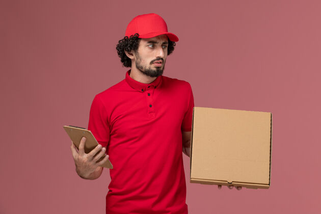 手持正面图：身穿红色衬衫和披风的男性快递员在粉色墙上拿着快递食品盒和记事本工作男服务