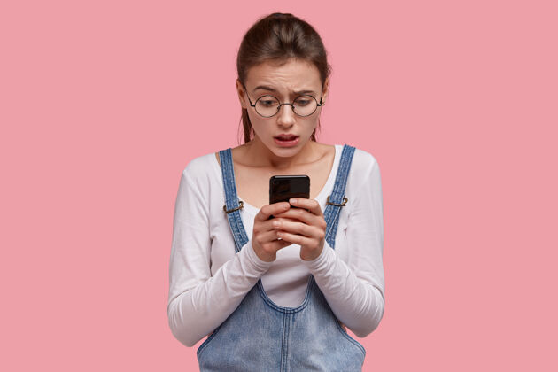 惊讶紧张不高兴的女人拿着现代手机 迷惑不解地在社交网络中收到不太喜欢她的帖子 穿着休闲服阅读智能手机手机