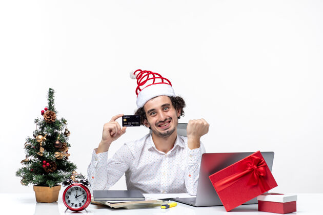 年轻年轻快乐的商人戴着圣诞老人的帽子 拿着他的银行卡 在白色背景的办公室里仔细地头脑风暴小心圣诞节商务人士