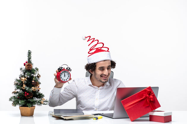 举行年轻搞笑情绪激动的商务人士戴着圣诞老人的帽子 拿着和展示的时钟坐在白色背景的办公室股票照片商务帽子坐着