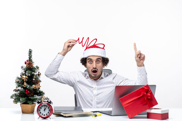 帽子新年的气氛与惊讶的年轻商人触摸他有趣的圣诞老人帽子指着上面的白色背景办公室圣诞老人商人圣诞老人