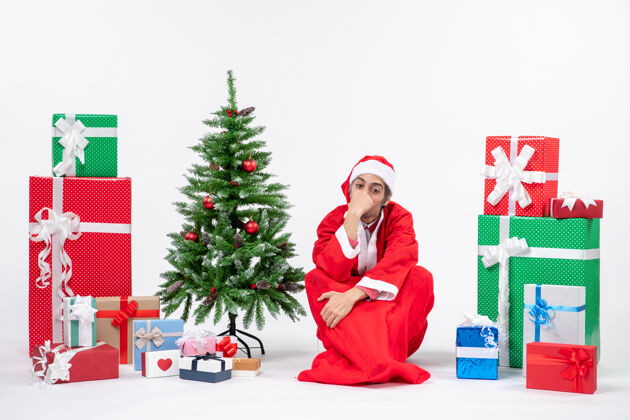 微笑悲伤体贴的年轻人打扮成圣诞老人 拿着礼物和装饰过的圣诞树坐在白色背景的地上圣诞老人礼物圣诞树