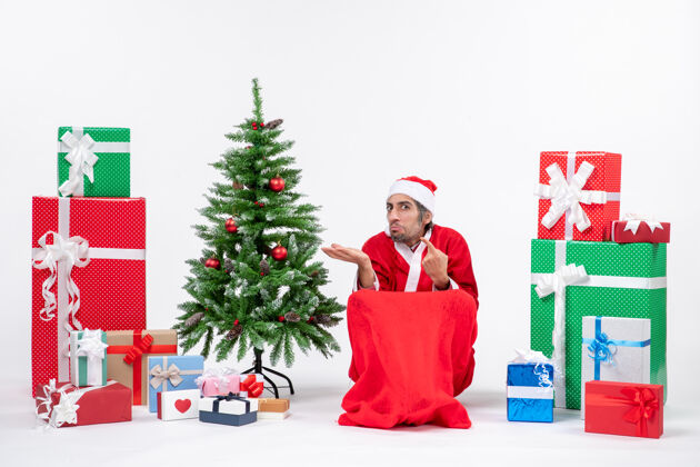 礼物好奇的年轻人打扮成圣诞老人与礼物和装饰圣诞树上的白色背景年轻卖家微笑