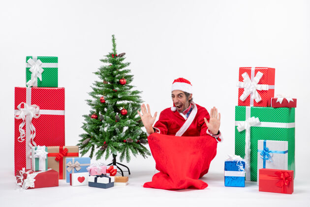 礼物骄傲的年轻人打扮成圣诞老人与礼物和装饰圣诞树坐在地上的白色背景人骄傲圣诞老人