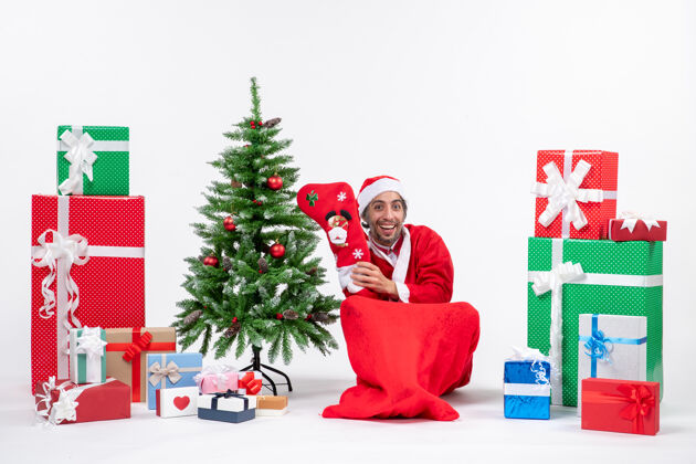 地积极的圣诞老人坐在地上 穿着圣诞袜靠近礼物和装饰在白色背景上的新年树礼物礼物衣服