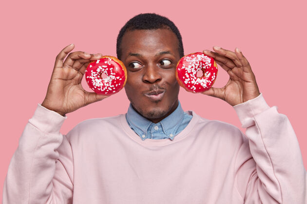 个人高兴的黑皮肤男性头像保持新鲜的甜甜圈靠近头部 穿着正式的衬衫和毛衣 模型在粉红色的空间非洲甜甜圈美国
