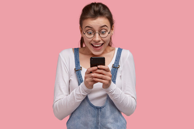牛仔高兴的欧洲年轻女子手持现代手机 凝视着通过互联网收到的令人敬畏的派对邀请 读着令人印象深刻的好消息电话智能手机短信