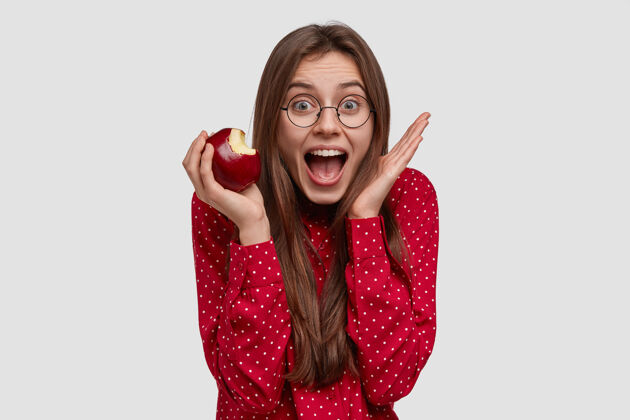 人类兴奋过度的快乐女士举手靠近脸 张大嘴巴 吃美味的苹果 戴透明眼镜饮食衬衫水果