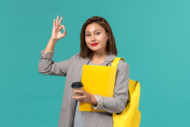 女学生身着灰色夹克 背着黄色背包 拿着文件 喝着咖啡的女学生在浅蓝色墙上微笑的正面照片背包夹克上课